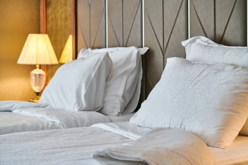 Two beds with white sheets in a bedroom with a lit lamp on the bedside table